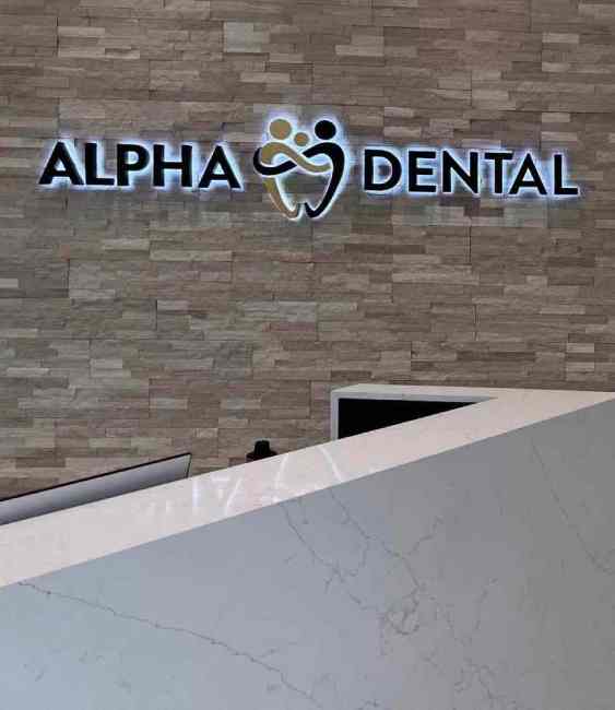 A picture of the Alpha Dental front desk with their logo displayed on the wall in the background. The reception area is clean and organized, with a modern desk and comfortable seating for patients. The logo on the wall represents the Alpha Dental brand.